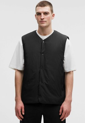 Vyras, dėvintis juodą užsegamą be rankovių liemenę ant baltos trumpomis rankovėmis palaidinės, stovintis priešais šviesų vienodą foną.