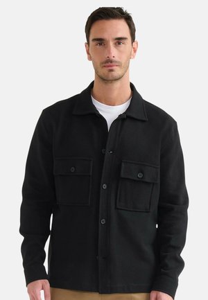 Giacca nera con bottoni e due tasche sul petto, realizzata in tessuto strutturato. Presenta un colletto e maniche lunghe, indossata da un modello.