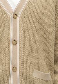 Μπεζ πλεκτή ζακέτα με κουμπιά από χελώνα, λευκή ριγέ τρέσα κατά μήκος της γραμμής κουμπιών και μικρή μπροστινή τσέπη.