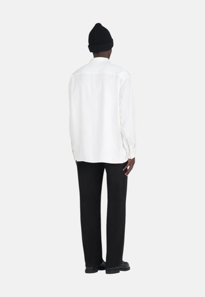 Chemise blanche à manches longues avec une coupe décontractée, épaulée de pantalons noirs. Le modèle porte également un bonnet en tricot noir et des chaussures noires.