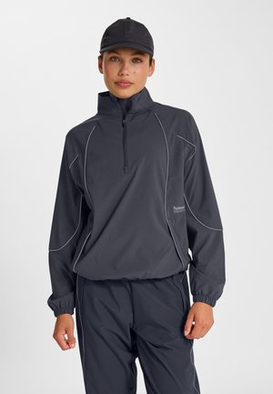 Vrouw in een atletisch jack van donkergrijs met een halve ritssluiting, bijpassende broek en zwarte pet, staand tegen een lichtgrijze achtergrond.