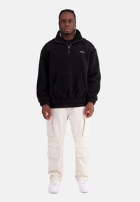 Schwarzer Fleece-Pullover mit Reißverschlusskragen und Fronttasche, kombiniert mit beigen Cargo-Hosen mit mehreren Taschen und weißen Sneakern.