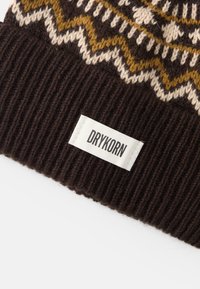 Καφέ πλεκτό σκουφί με ραβδωτή υφή, που διαθέτει γεωμετρικό μοτίβο σε κρεμ και χρυσό. Περιέχει λευκή ετικέτα με την επιγραφή "DRYKORN".