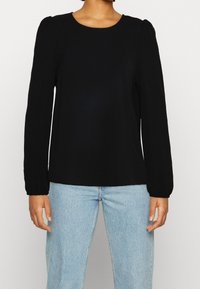 Maglietta nera a maniche lunghe con scollo rotondo, caratterizzata da dettagli arricciati sulle spalle e polsini elasticizzati, abbinata a jeans azzurri chiari.