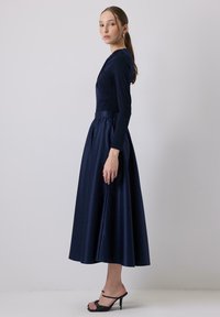 Tengerészkék, hosszú ujjú ruha, illeszkedő felső résszel és bővülő szoknyával. Simább szövet, amely finom fényt kapott, hangsúlyozott derékvonallal és minimalista dizájnnal.