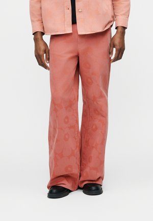 Mann trägt locker sitzende, rosafarbene Hose mit dezenten Blumenmustern und schwarzen Schuhen, steht vor einem schlichten Hintergrund.