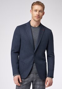 Blazer blu navy in tessuto testurizzato, a un petto con due bottoni, abbinato a una maglietta grigia a girocollo e pantaloni grigi a quadri.