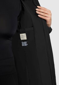 Kobieta w czarnym stroju trzyma otwartą czarną marynarkę, ukazując dwie wewnętrzne metki z nazwą marki.