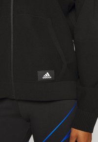 Chaqueta negra de Adidas con un tejido de punto texturizado, que cuenta con una cremallera, bolsillos laterales y un parche con el logo en la parte inferior izquierda.