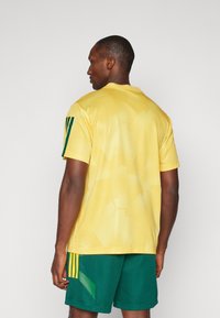 Κίτρινη αθλητική μπλούζα από διχτυωτό ύφασμα με πράσινες λεπτομέρειες και ρίγες στα μανίκια, σε συνδυασμό με πράσινες υφασμάτινες βερμούδες. Διαθέτει ribbed γιακά και αθλητικό σχέδιο.