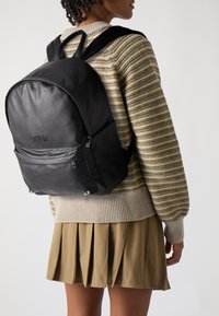 Mochila de cuero negro con forma redondeada, bolsillo delantero con cremallera y herrajes plateados. Persona vistiendo un suéter a rayas y una falda plisada.