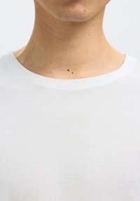 Vrat i donji dio lica osobe koja nosi bijelu košulju, s dvije istaknute tamne madeže na prednjem dijelu vrata.
