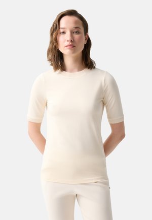 Jeune femme aux cheveux bruns ondulés portant une chemise crème à manches courtes et un pantalon assorti, debout, les mains derrière le dos, sur fond blanc.