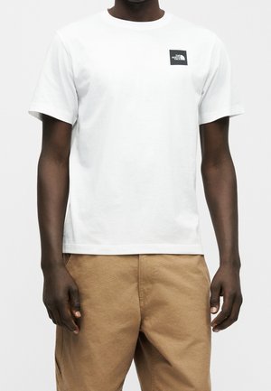 Homme portant un t-shirt blanc uni à manches courtes avec un petit logo carré noir sur la poitrine gauche et un pantalon kaki.