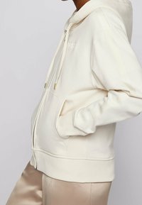 Sudadera con cremallera en crema, fabricada en un tejido suave, con cordones y detalles en dorado. Presenta un bolsillo tipo canguro y puños acanalados. Diseño sencillo, sin patrones.