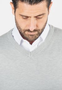 Pull léger gris clair, en V, composé d'un tissu tricoté par-dessus une chemise blanche à col. Il présente des poignets côtelés et une finition soignée.