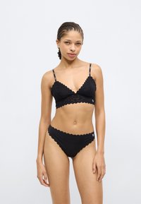 Juoda nėriniuota liemenėlė su žvyneliais krašteliais ir reguliuojamais dirželiais, derinama su atitinkamomis bikinio kelnaitėmis su žvyneliais krašteliais, abi turi lygią tekstūrą.