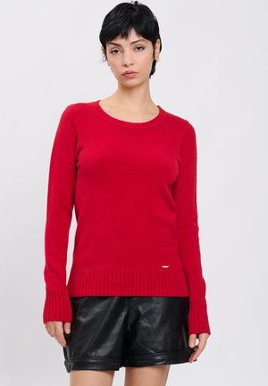 Kvinde med kort sort hår iført en rød langærmet sweater og sorte lædershorts står mod en ensfarvet baggrund.