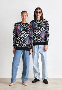 To modeller står side om side iført svarte hettegensere med fargerike mønstre, hvit tekst, lyseblå jeans og nøytralt fottøy.
