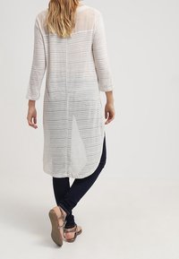 Dlouhá světle šedá tunika s vodorovnými pruhy, průsvitným živůtkem a rozparkem vzadu, doplněná tmavě modrými legínami a hnědými sandály.