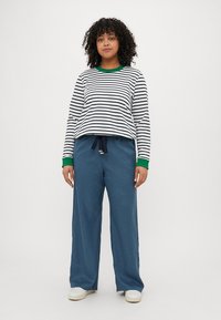 Maglietta a righe a maniche lunghe bianca e navy con polsini verdi, abbinata a pantaloni blu larghi e sneaker bianche. Outfit casual con vestibilità rilassata.