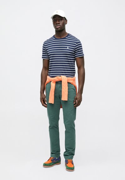 Μπλε μπλουζάκι με κοντά μανίκια και ρίγες, πορτοκαλί πουλόβερ δεμένο γύρω από τη μέση, πράσινο παντελόνι και πολύχρωμα αθλητικά παπούτσια με καφέ σόλες.