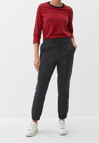 Chemise à manches longues rayée rouge et noire, associée à un pantalon gris foncé effilé avec poches et poignets élastiques, portée avec des baskets blanches.