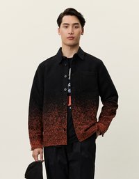 Fekete és narancssárga árnyalatú ingdzseki gombos záródással, egyetlen mellzsebbel és struktúrált anyaggal, fekete grafikázott póló felett viselve.