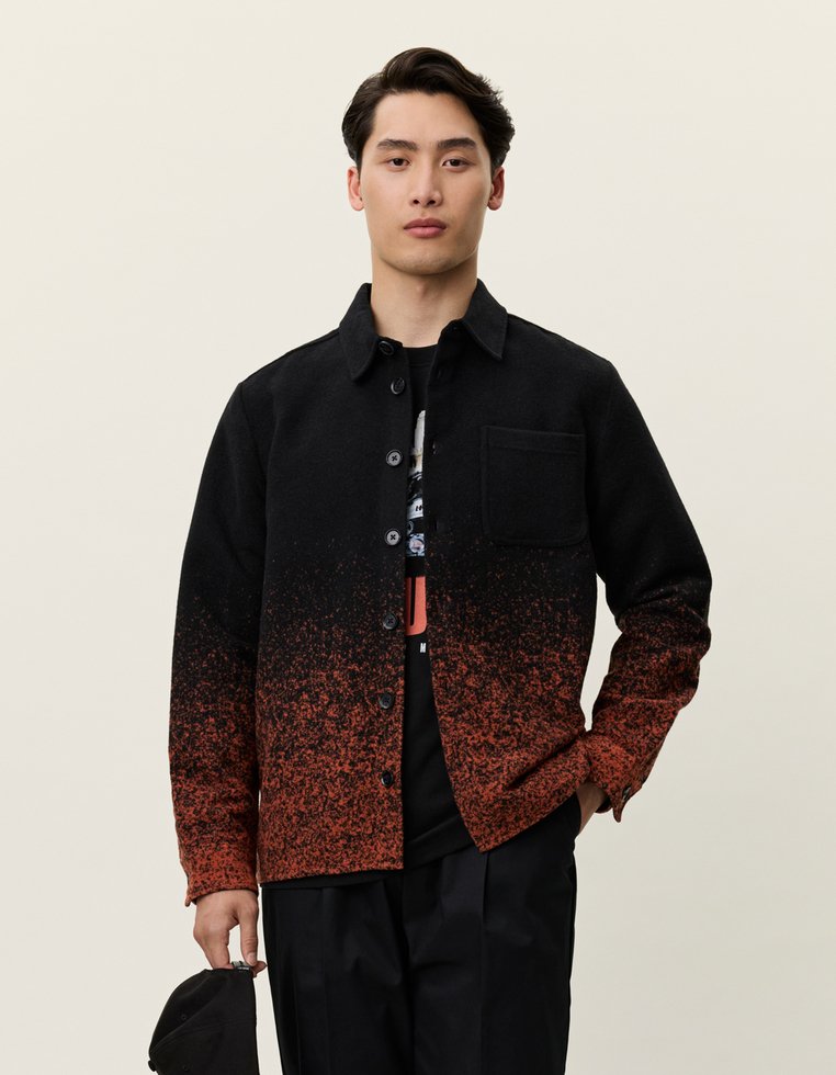 Fekete és narancssárga árnyalatú ingdzseki gombos záródással, egyetlen mellzsebbel és struktúrált anyaggal, fekete grafikázott póló felett viselve.