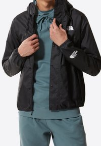 Homme portant un sweat à capuche bleu sarcelle et un pantalon assorti, ajustant une veste noire The North Face avec des logos sur la poitrine et la manche.