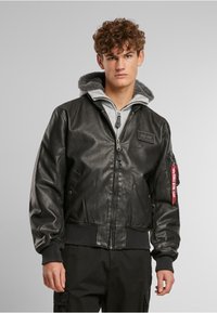 Veste bomber en cuir noir avec capuche doublée en polaire grise, poignets et ourlet côtelés, fermeture éclair frontale et étiquette en tissu rouge sur la manche.