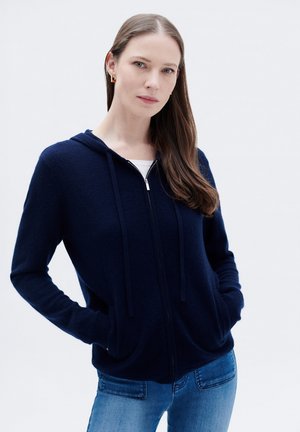 Haut à zip bleu marine en tissu doux, avec une capuche, des cordons de serrage, des poches latérales et des poignets côtelés. Associé à un jean bleu clair.