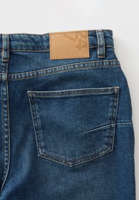 Jeans van denim in een donkerblauwe kleur, met een gestructureerde stof en een tan leren patch op de taille met een reliëflogo en detail op de zak.