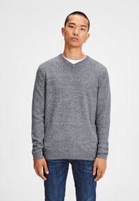 Grauer V-Ausschnitt-Pullover aus strukturiertem Strickstoff, mit langen Ärmeln und einem figurbetonten Saum, kombiniert mit dunkelblauen Jeans.