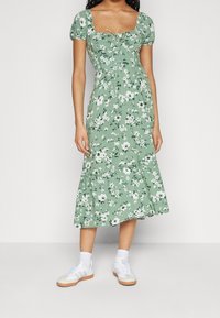 Zielona sukienka w kwiaty z bufkami na rękawach, kwadratowym dekoltem i marszczonym pasem. Stworzona z białymi trampkami i skarpetkami do kostek.