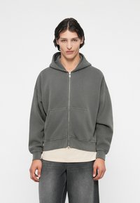 Harmaa vetoketjuhuppari, jossa on nyörikiinnitys, etutaskut, löysä malli ja ribbikuvioidut hihansuuts. Päällä vaaleanvärinen paita ja harmaat farkut.