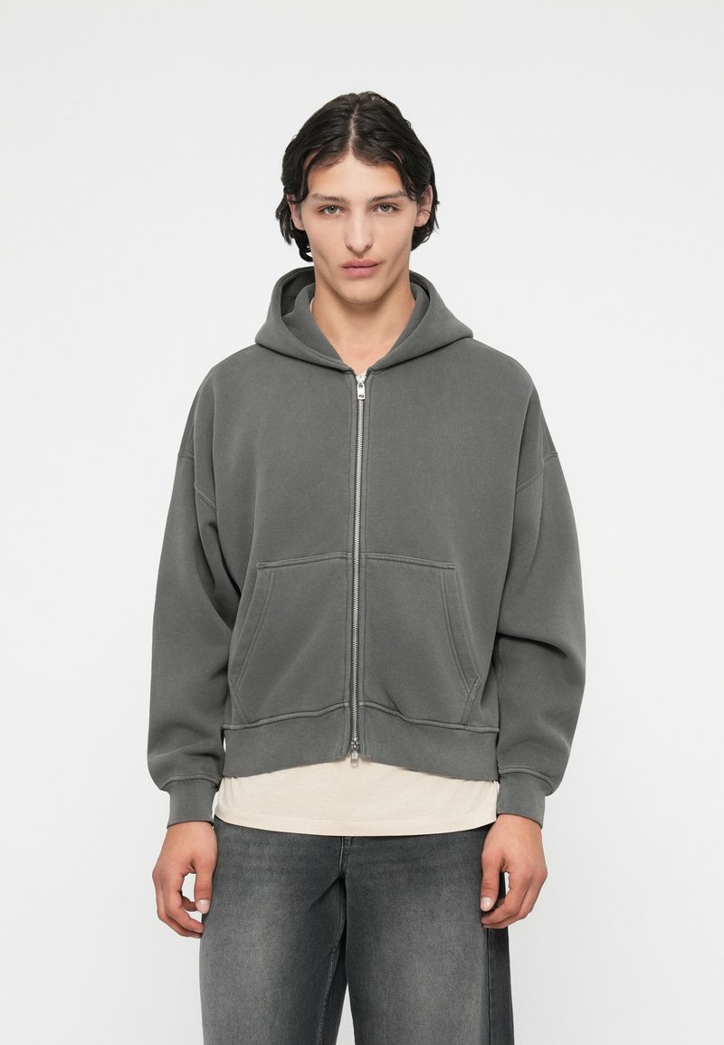 Harmaa vetoketjuhuppari, jossa on nyörikiinnitys, etutaskut, löysä malli ja ribbikuvioidut hihansuuts. Päällä vaaleanvärinen paita ja harmaat farkut.