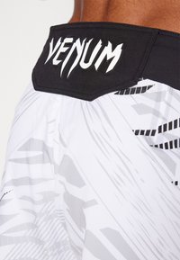 Bele športne kratke hlače z črnimi pasom, na katerih je v beli barvi logo "VENUM". Na hlačah so vidni abstraktni sivi vzorci.