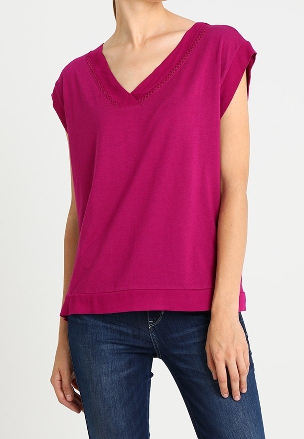 Vrouw draagt een mouwloze magenta top met V-hals en donkerblauwe spijkerbroek, staand tegen een witte achtergrond.