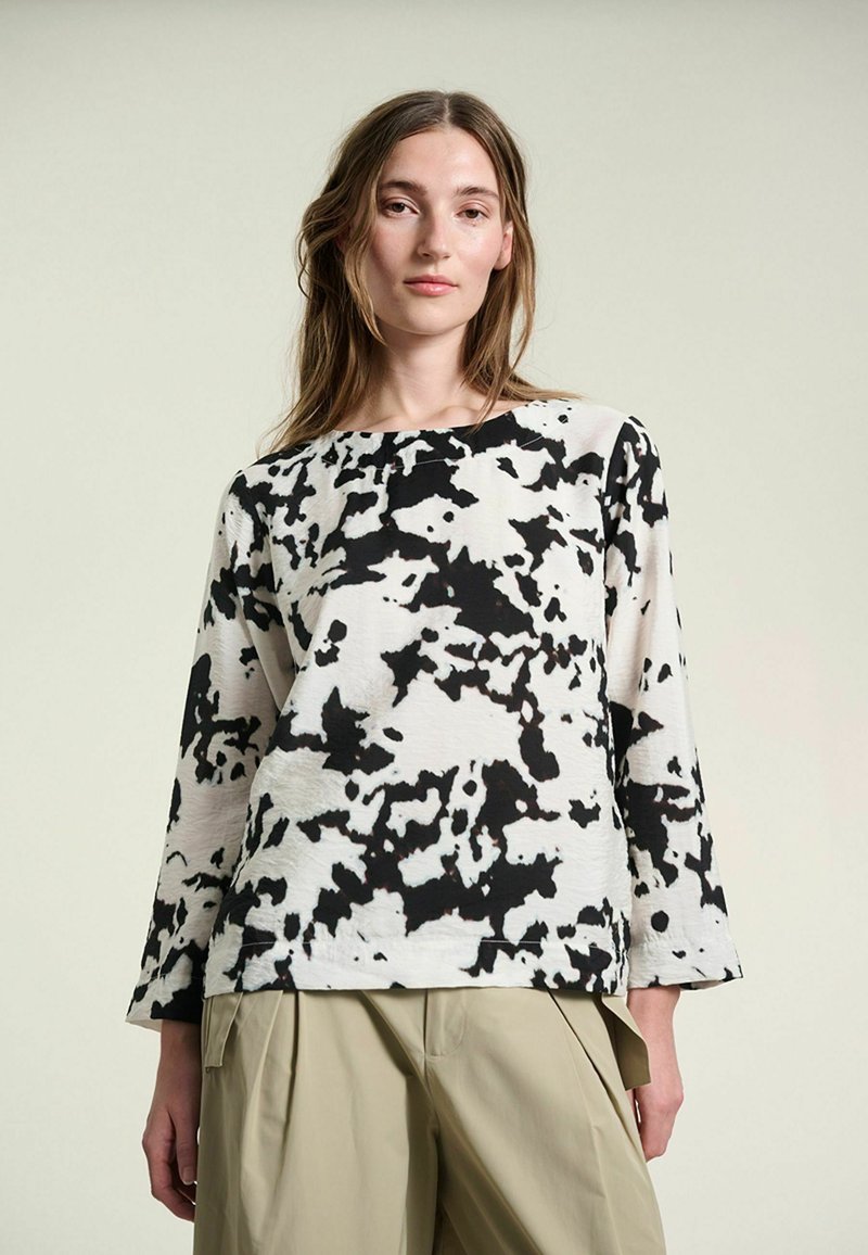 Jeune femme portant un haut à manches longues à motif abstrait noir et blanc et un pantalon beige taille haute à jambes larges, debout devant un fond uni.