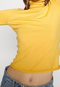 Maglione giallo a collo alto con una vestibilità aderente e una texture morbida, abbinato a jeans in denim blu strappati. Presenta una lunghezza cropped.