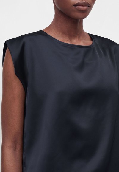 Gros plan d'une personne portant un top sans manches bleu marine foncé en satin avec un col rond sur un fond neutre.