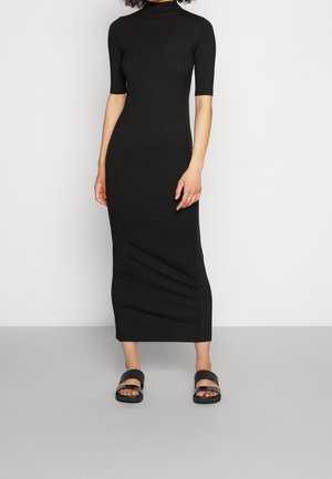 Kobieta ubrana w dopasowaną, czarną, prążkowaną sukienkę midi z rękawami do łokcia oraz czarne klapki, stojąca na tle jednolitego tła.