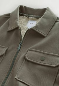 Brown corduroy jacket with a zip closure, fleece lining, two chest pockets, and a collared neck. Tag visible inside with brand name.