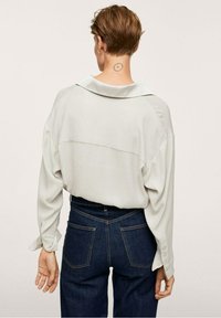 Henkilö, jolla on lyhyet ruskeat hiukset, pukeutuu löysään vaaleanharmaaseen paitaan ja tummansinisiin farkkuihin, ja heillä on ympyrämainonta kaulan takana.