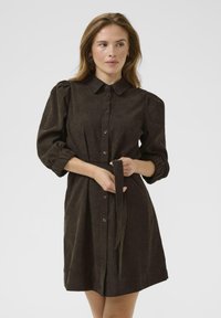 Robe brune à boutons avec un col, des manches bouffantes et une ceinture assortie. Fabriquée en tissu texturé, elle présente une coupe décontractée et une longueur mi-genou.