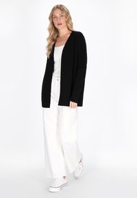 Cardigan nero a coste in morbido knit, design aperto davanti, con maniche lunghe. Abbinato a una maglietta bianca e pantaloni bianchi larghi, e sneakers bianche.