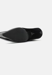 Zapato de tacón alto de charol negro con punta afilada y suela texturizada. El tacón presenta un acabado liso y un diseño elegante y aerodinámico.