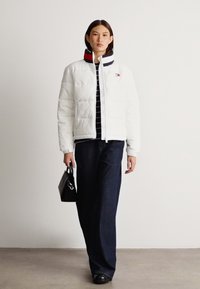 Balta pūkinė striukė su siuvinėta tekstūra, raibu apykakle su raudonais ir tamsiai mėlynais akcentais, derinama su tamsiai mėlynomis džinsinėmis kelnėmis ir juoda rankine.