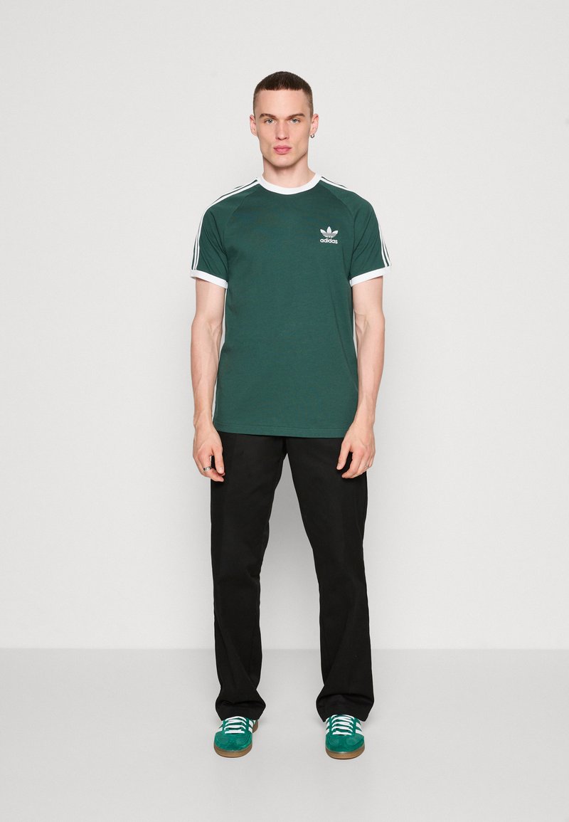 T-shirt en coton vert avec col et manches côtelés blancs, arborant un petit logo brodé sur la poitrine, associé à un pantalon noir et des chaussures vertes.
